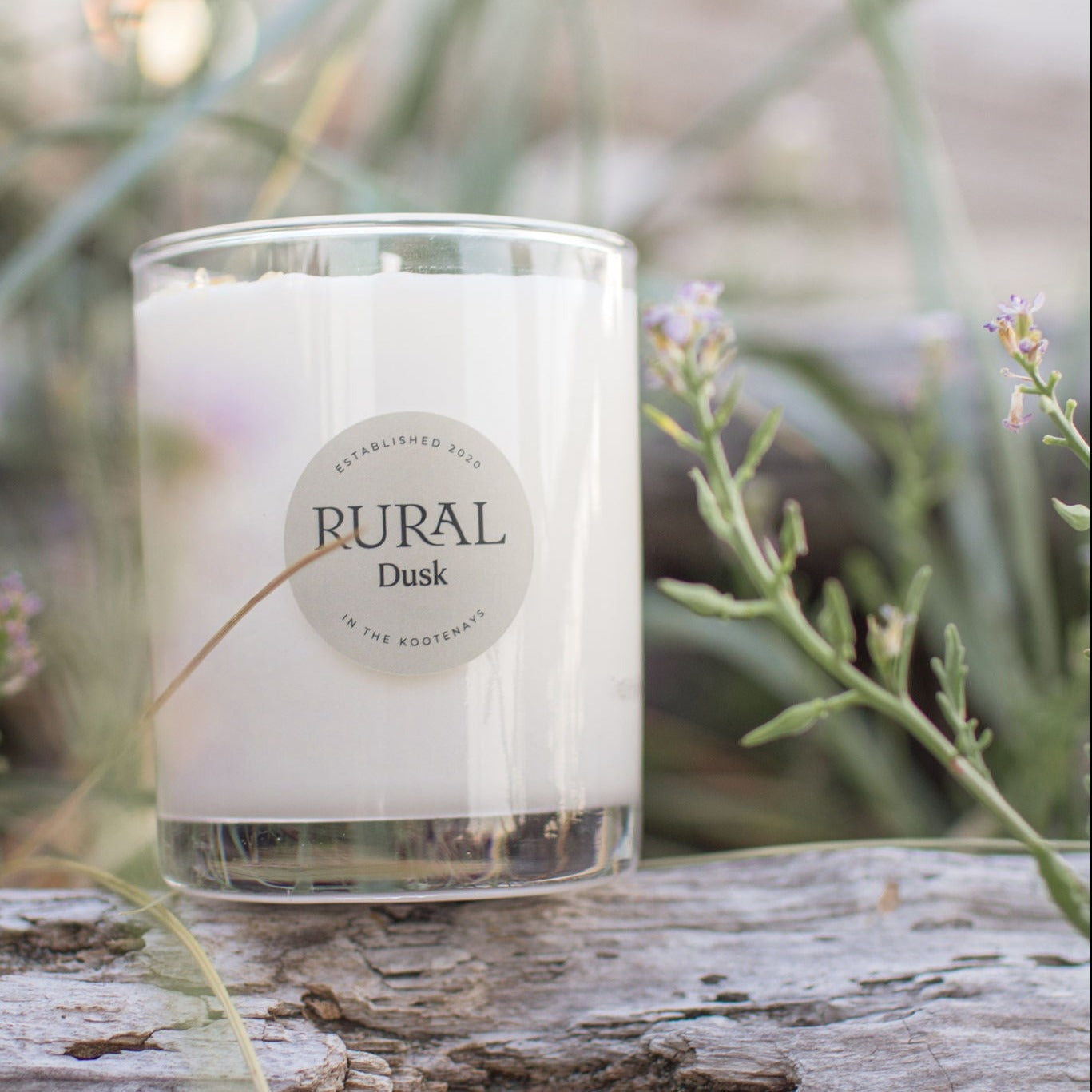 Photo is of a white candle in a clear glass with a round tan colored label indicating RURAL Candles Dusk scent. The candle is sitting on a driftwood log and surrounded by wild flowers and tall grasses.