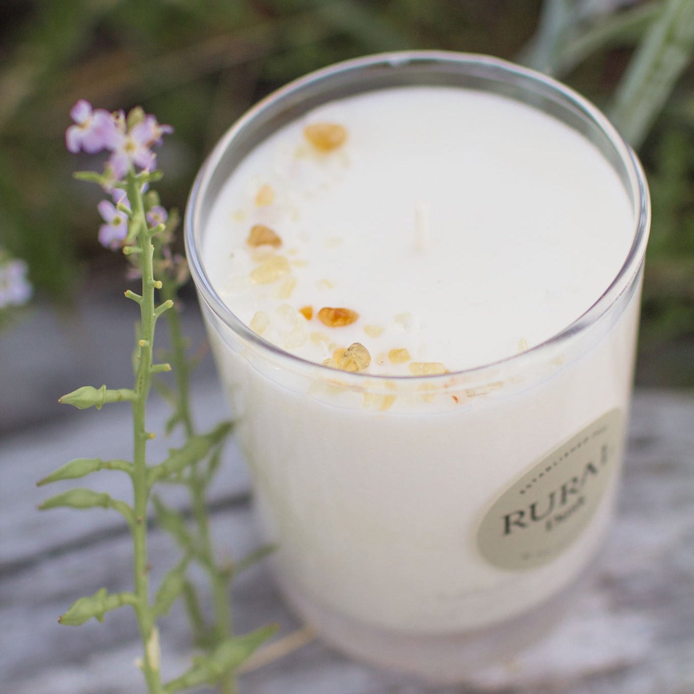Top view photo of a white candle in a clear glass with a round tan colored label indicating RURAL Candles Dusk scent. The candle is sitting on a driftwood log and surrounded by wildflowers and tall grasses. We can see the top of the candle is sprinkled with golden citrine gemstones in a crescent moon shape.