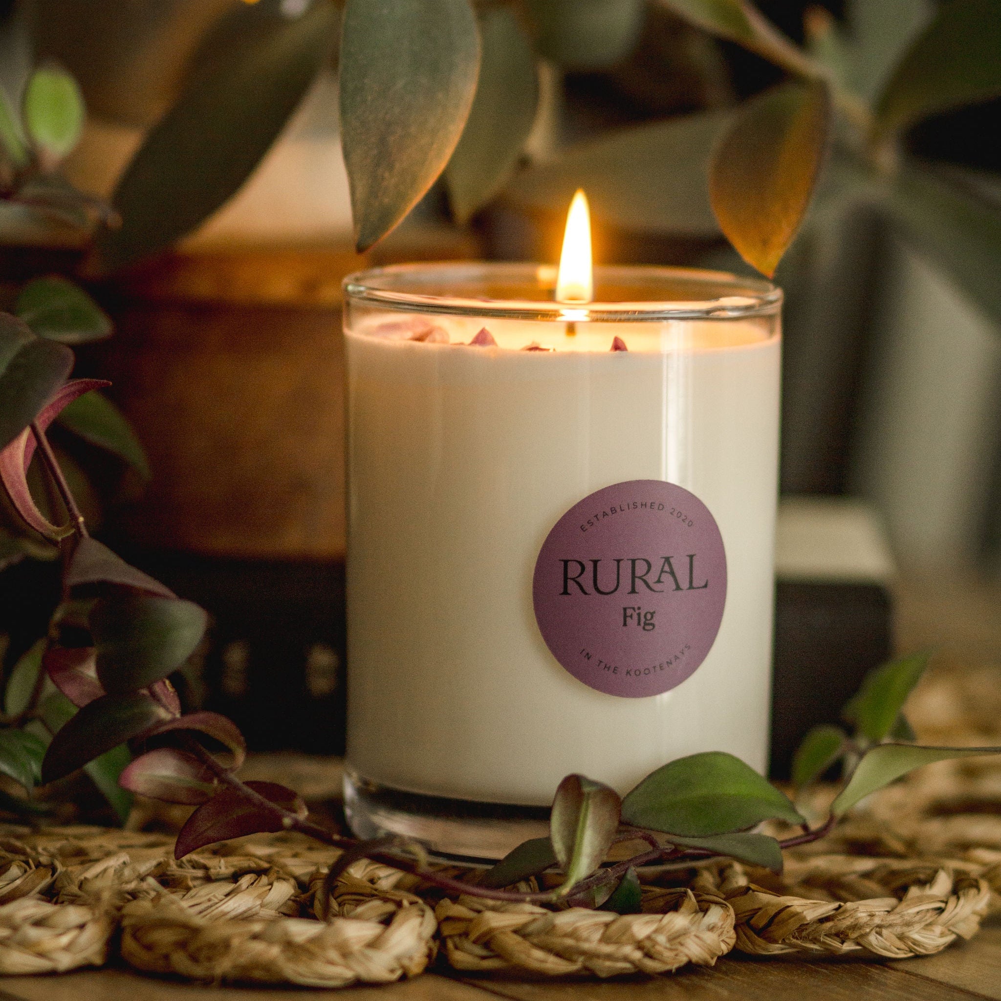 Candle with a 'Rural' label on a wooden surface with plants in the background