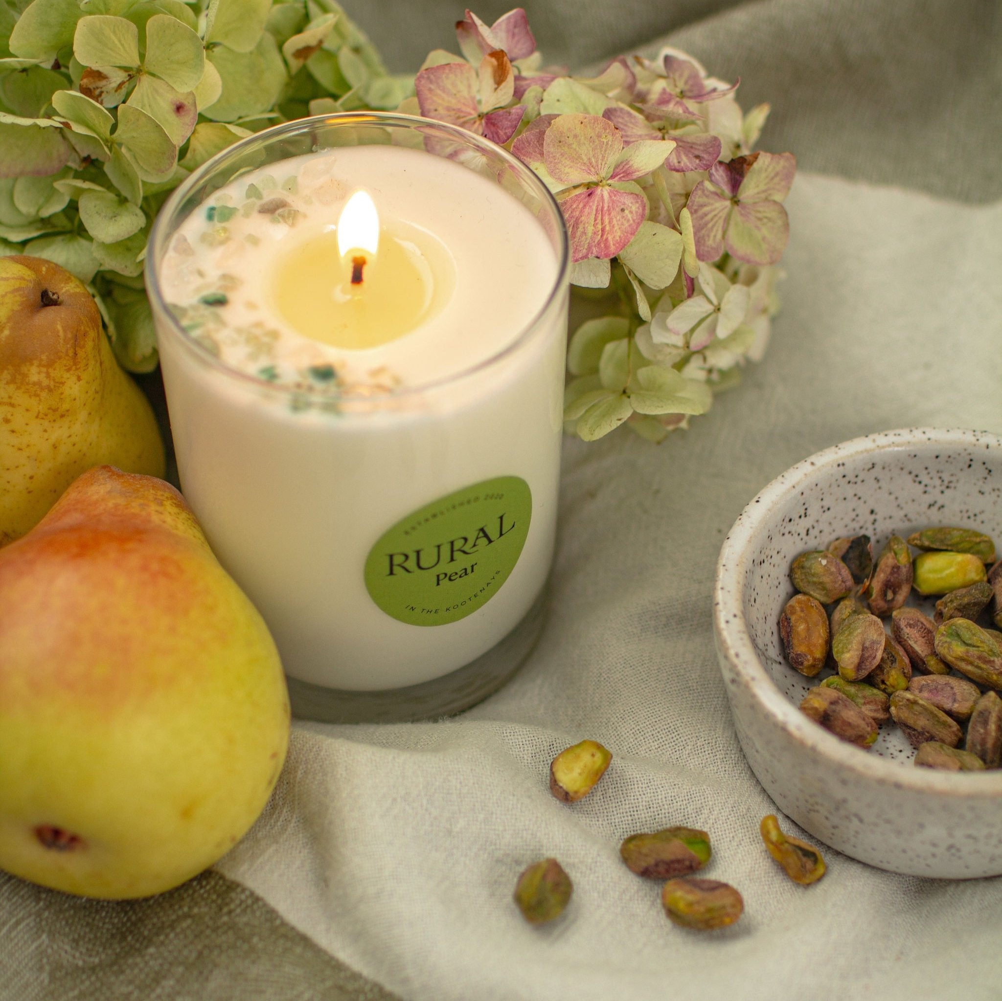 Photo of a lit white candle in a clear glass with a round green label indicating RURAL Candles Pear Scent. We can see the top of the candle is decorated with small green gemstones, artfully arranged in a crescent moon shape. The candle is surrounded by fresh pears, pistachios and flowers on a linen cloth, very fresh and inviting kitchen scene.