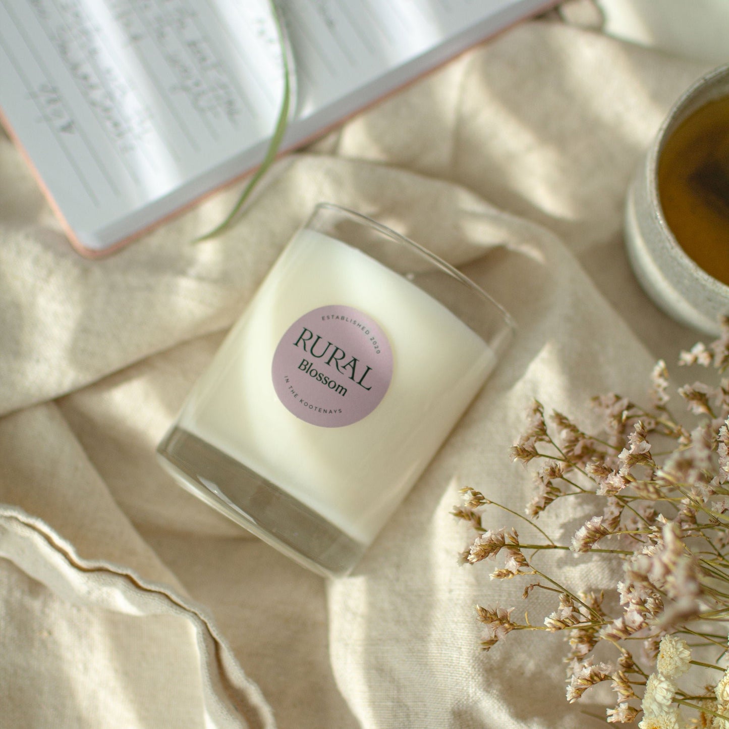 Photo of a white candle in a classic glass container with a light purple round RURAL Candles label on it, indicating the scent is called Blossom. The mood of the photo is very dreamy and feminine, with flowers, linen, tea, and a handwritten journal in the background.