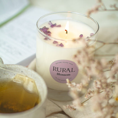 Photo of a white candle in a clear glass container with a light purple round RURAL Candles label on it, indicating the scent is called Blossom. The candle top is embellished with a sprinkling of purple amethyst chips, and the candle is sitting on a table linen with tea, wildflowers and a handwritten journal in the background.