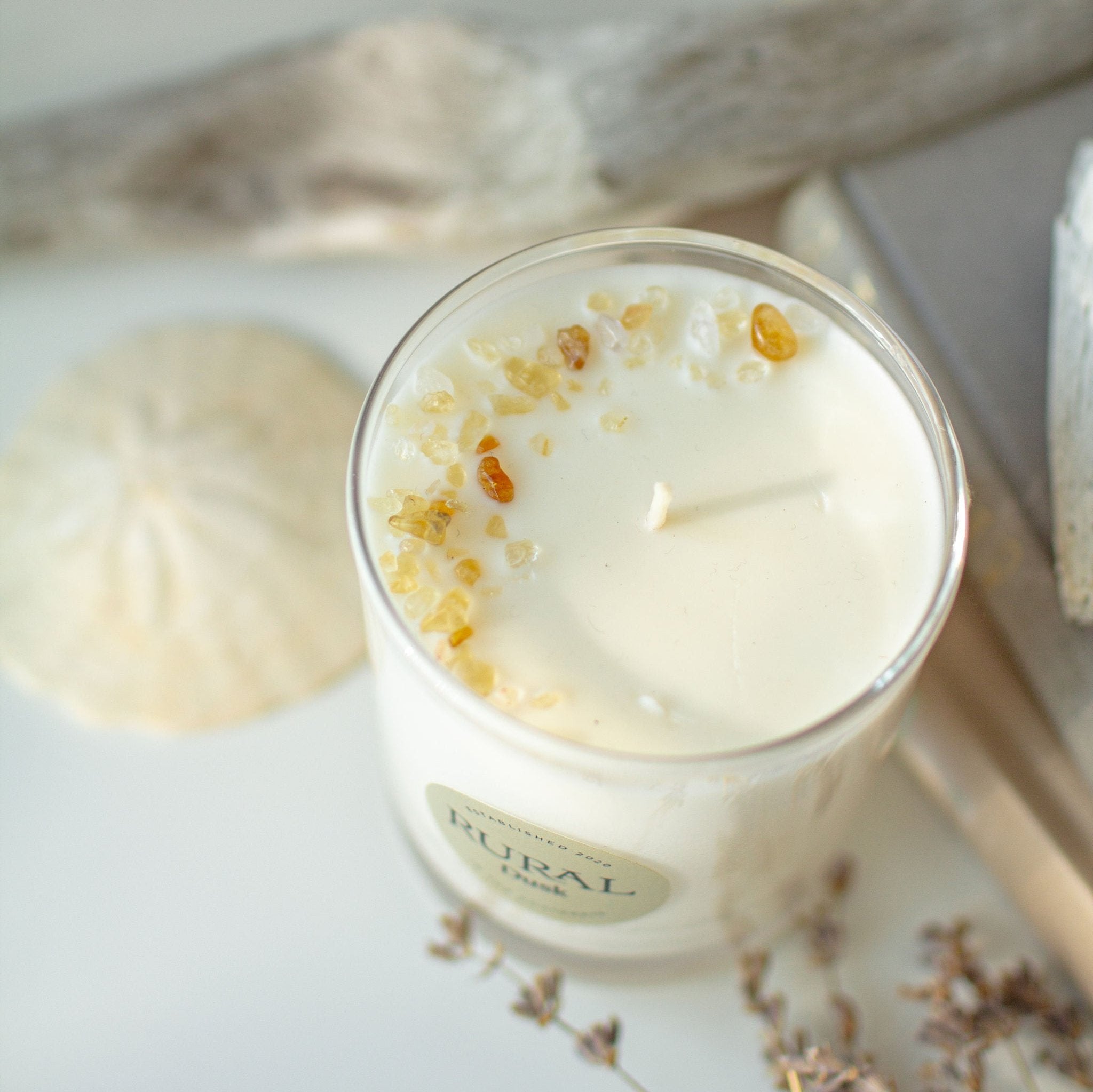 Top view photo of a white candle in a clear glass container. The top of the candle is decorated with golden citrine gemstones, arranged in a crescent moon shape. Seashells, driftwood and flowers are in the background, creating a serene, coastal vibe.