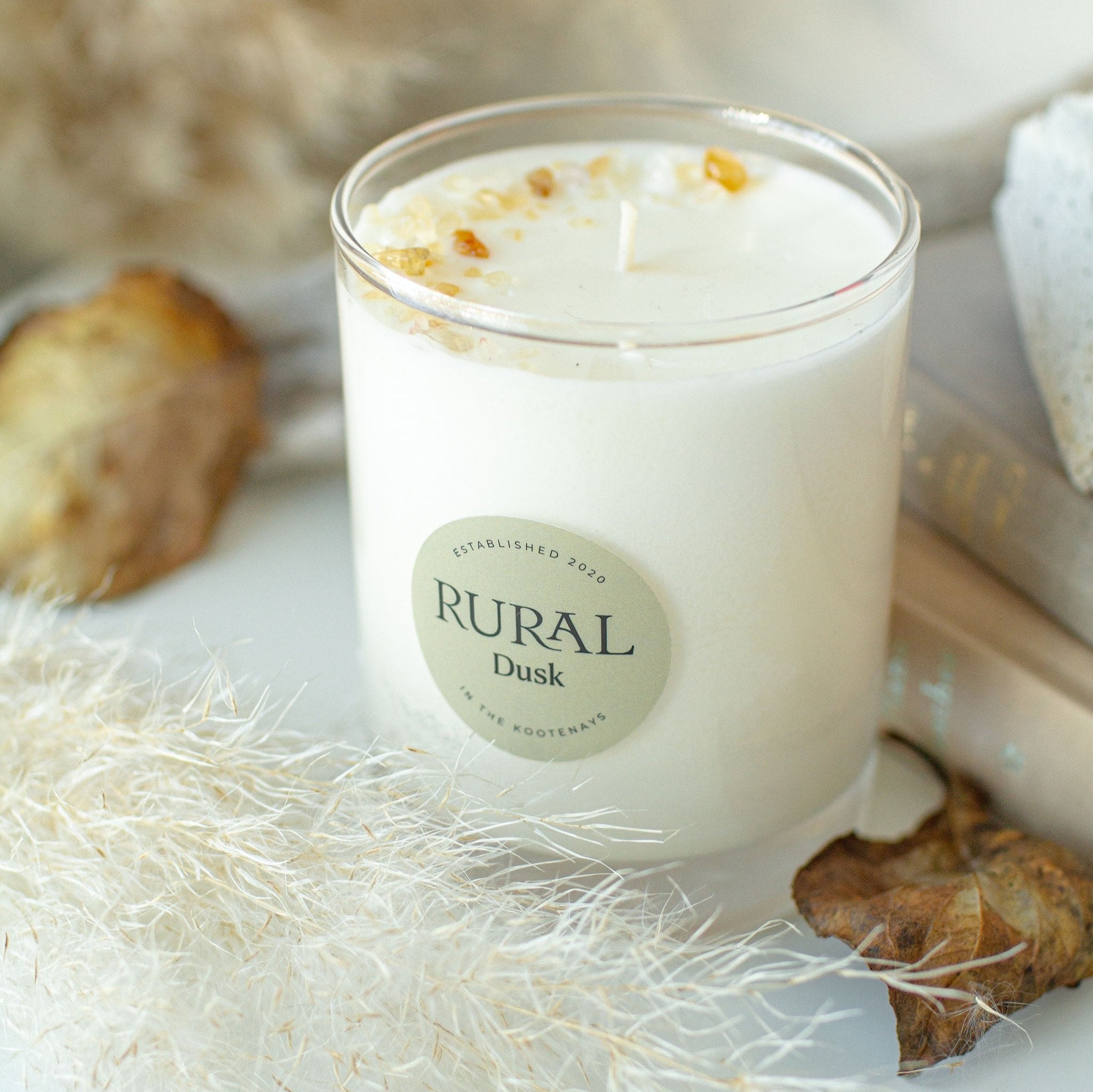 Photo of a Dusk scented RURAL Candle in a clear glass. The candle is  sitting on a linen cloth with dried leaves, flowers and books surrounding it, creating a cozy Fall morning bedside or in a living room.