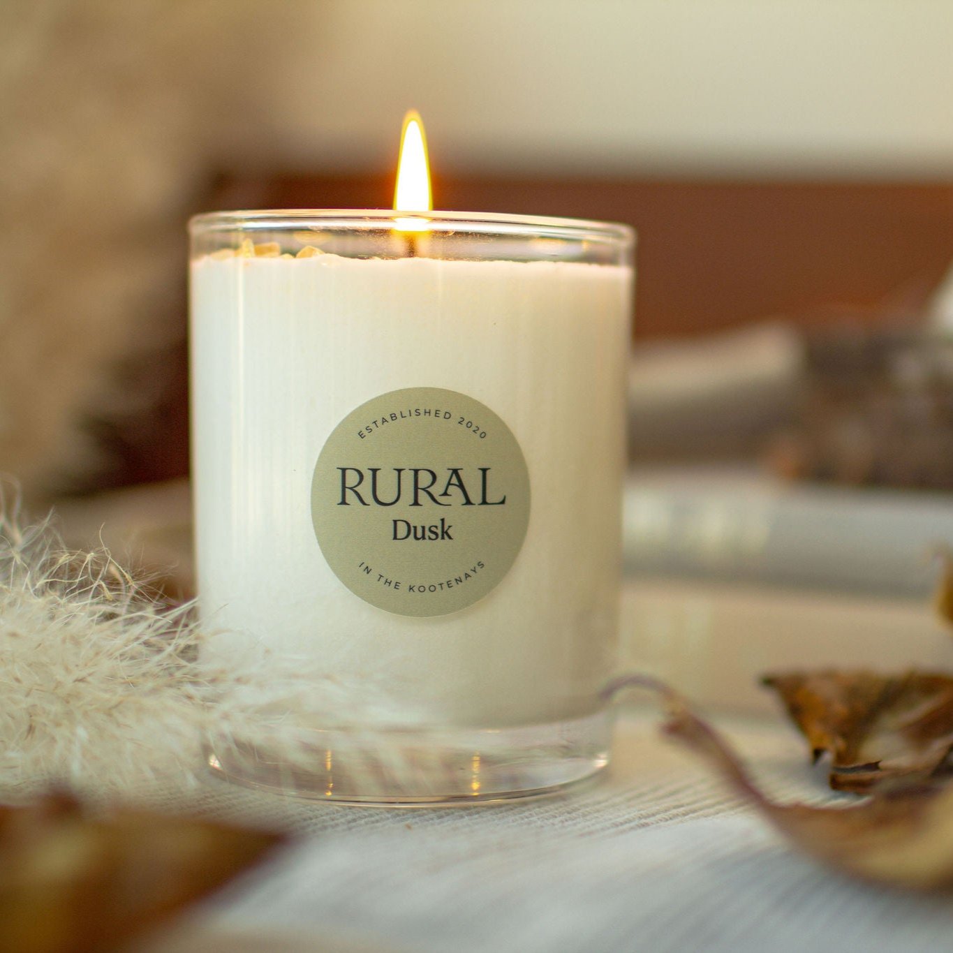 Photo of a Dusk scented RURAL Candle in a clear glass. The candle is lit and sitting on a linen cloth with dried leaves and flowers surrounding it, creating a cozy Fall evening.