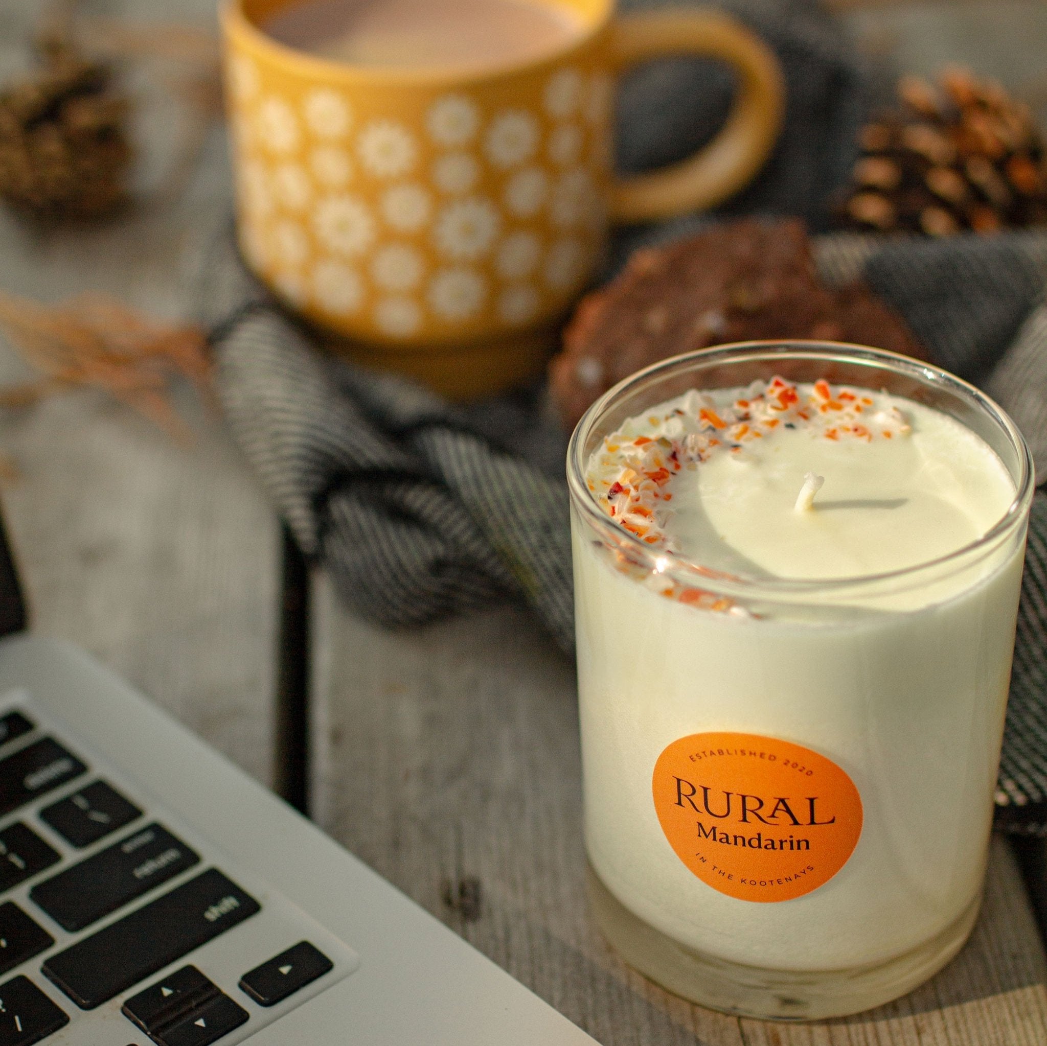 Photo of a RURAL Candles Mandarin scented candle in a clear glass with a round orange label. We can see the top of the candle is decorated with small orange carnelian gemstones and clear quartz crystals, artfully arranged in a crescent moon shape. The candle is on a wooden desk with pinecones, wool blanket, a mug of tea and a computer, for a cozy Fall home office space.