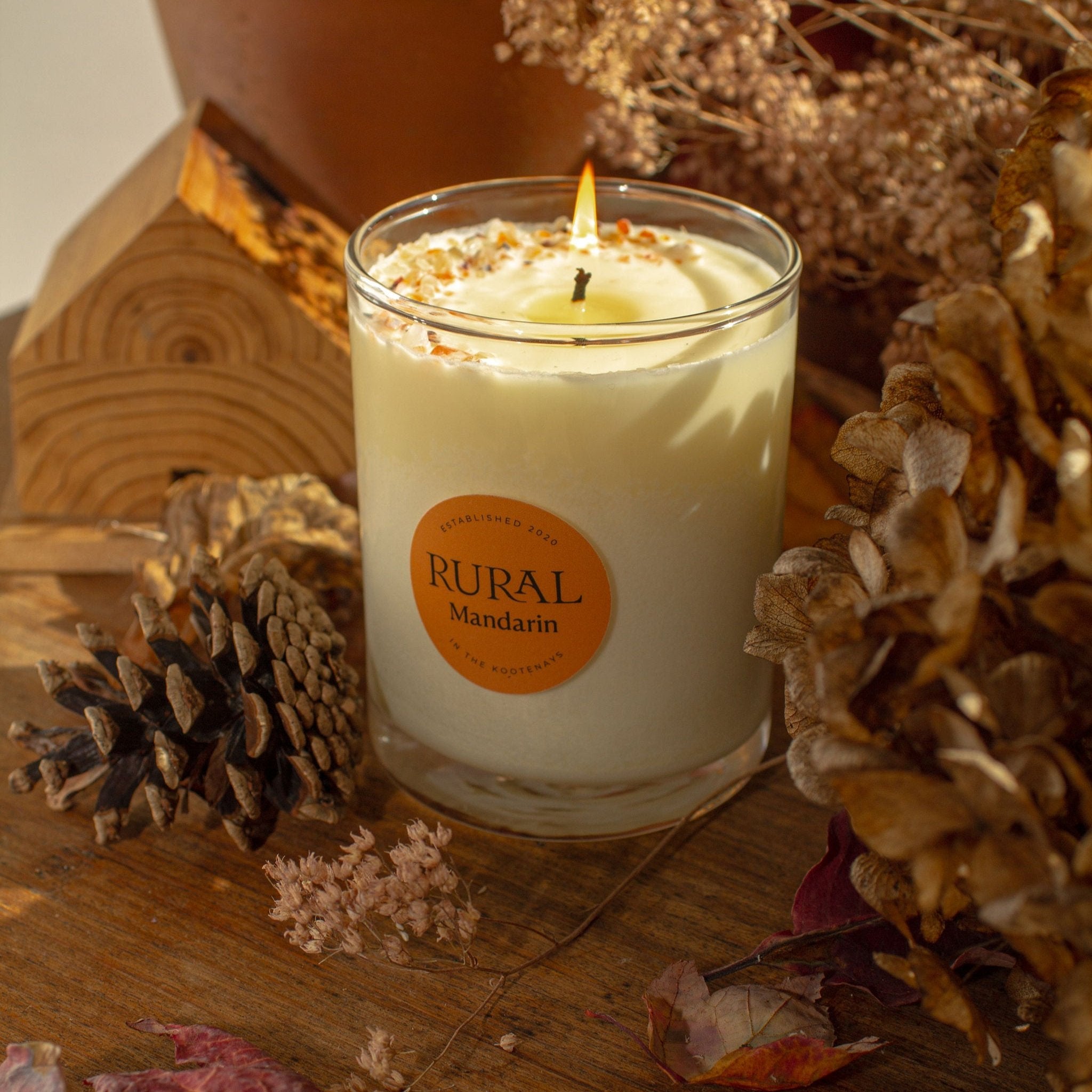 Photo of a RURAL Candles Mandarin scented candle in a clear glass with a round orange label. We can see the top of the candle is decorated with small orange carnelian gemstones and clear quartz crystals, artfully arranged in a crescent moon shape. The candle has a gentle flame and is on a wooden table with pinecones and dried flower decor for a cozy Fall feel.