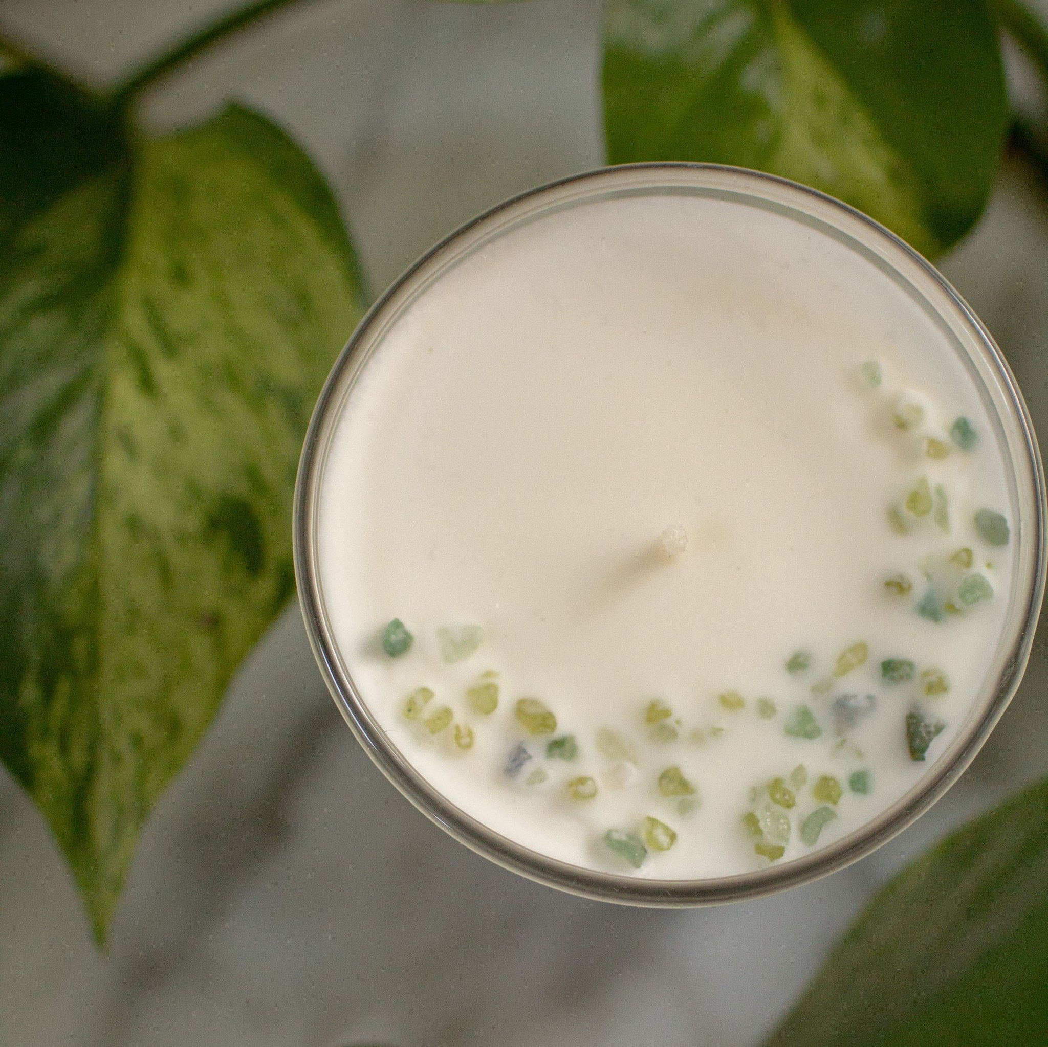 Top view photo of a coconut soy candle in a clear white glass, sitting on a marble counter with green leaves surrounding it, for a fresh vibe and inviting space. We can see the top of the candle is decorated with little green gemstones, artfully arranged in the shape of a crescent moon.