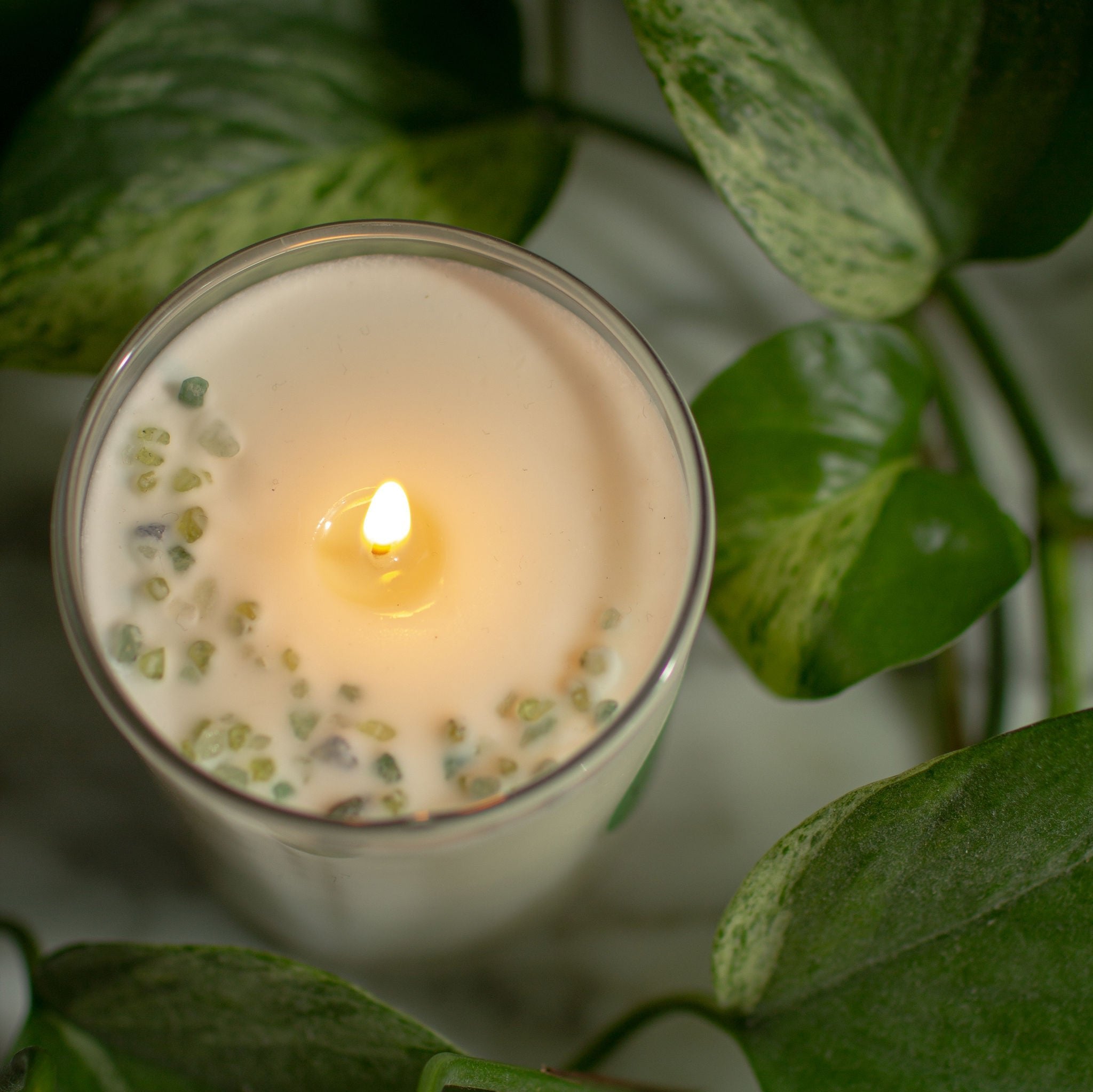 Top view photo of a coconut soy candle in a clear white glass, this time it's lit, sitting on a marble counter with green leaves surrounding it, for a serene vibe and lush, inviting space. We can see the top of the candle is decorated with little green gemstones, artfully arranged in the shape of a crescent moon.