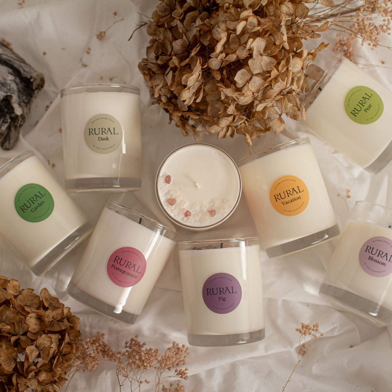 Group photo of coconut soy candles in clear glass with gemstones on top and colorful round labels indicating RURAL scented candles in Garden, Dusk, Pomegranate, Fig, Vacation, Pear, and Blossom fragrances.