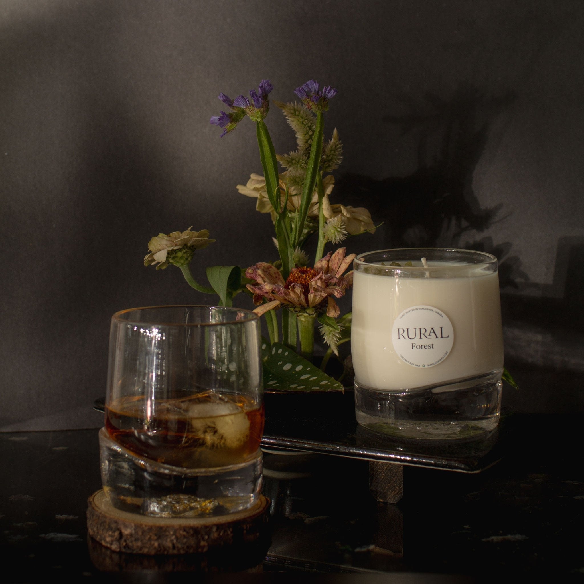 The photo is a moody, mysterious compilation of two very unique hand-blown, heavy-bottom glass whiskey tumblers. One has been handcrafted into a RURAL Candles Forest scented candle, and the other holds a drink, could be whiskey on the rocks. In the background is an exotic bouquet of flowers.