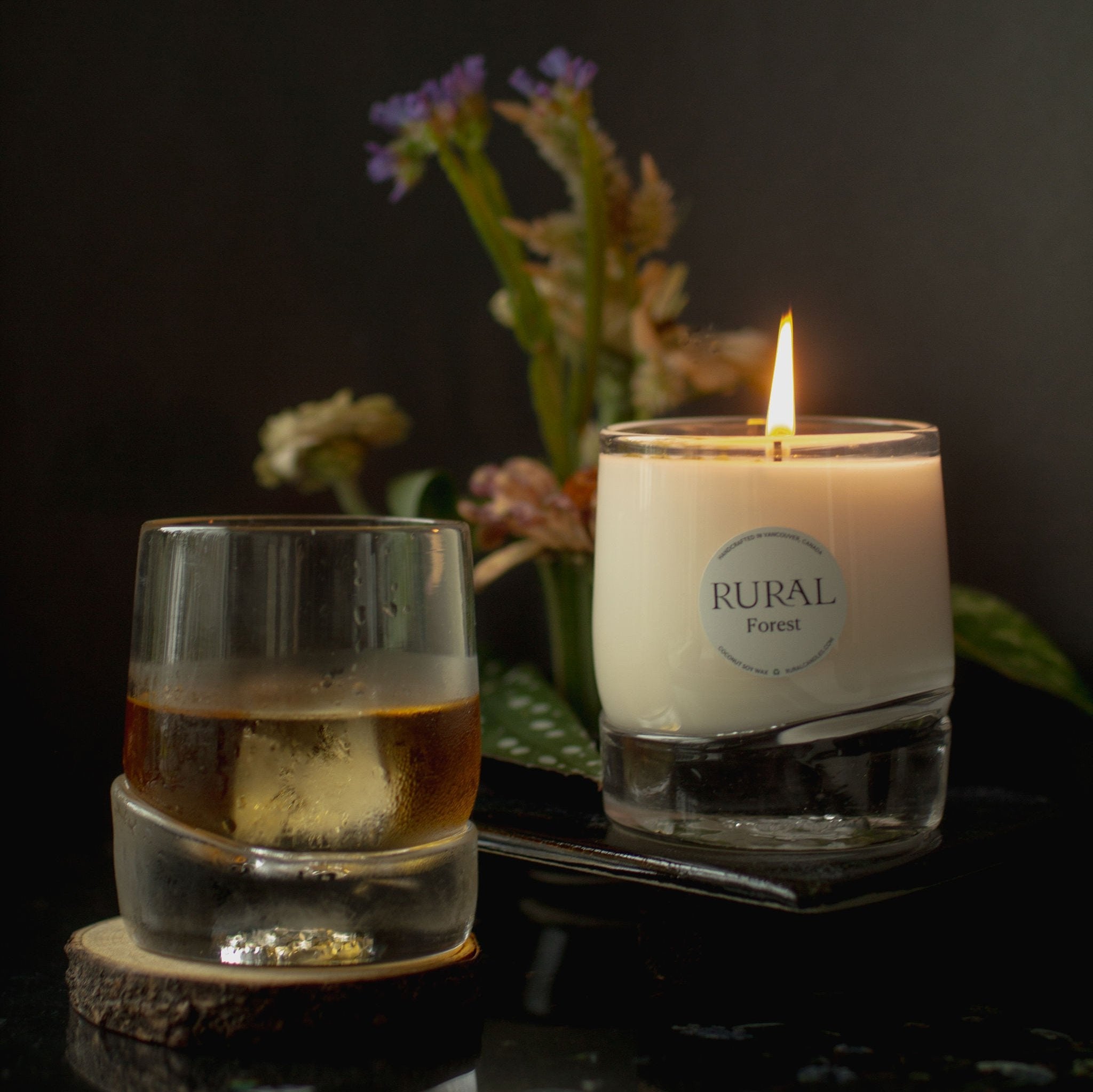 Photo of two very unique hand-blown, heavy-bottom glass whiskey tumblers. One has been handcrafted into a RURAL Candles Forest scented candle, and the other holds a drink, which could be whiskey on the rocks. In the background is an exotic bouquet of flowers and natural wood and black leather decor. The candle is lit, providing a warm and inviting cocktail hour mood.