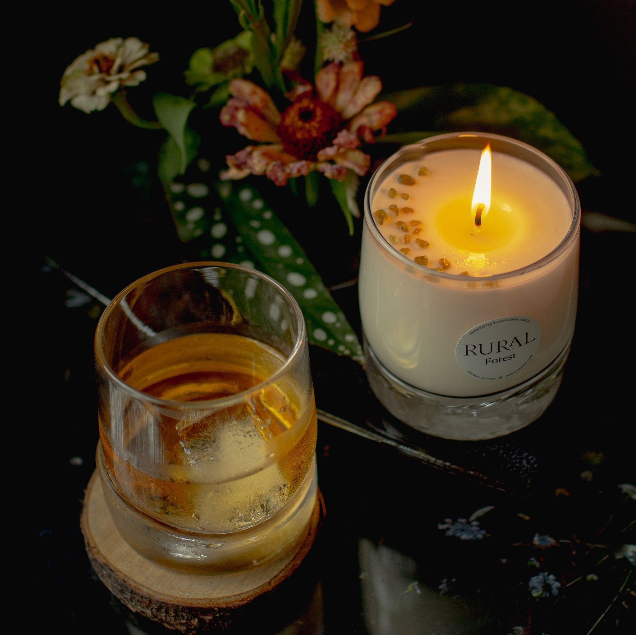 Top view photo of two very unique hand-blown, heavy-bottom glass whiskey tumblers. One has been handcrafted into a RURAL Candles Forest scented candle with gemstones on top, and the other holds a drink, could be whiskey on the rocks. In the background is an exotic bouquet of flowers. The candle is lit, providing a warm and inviting, cocktail hour mood.