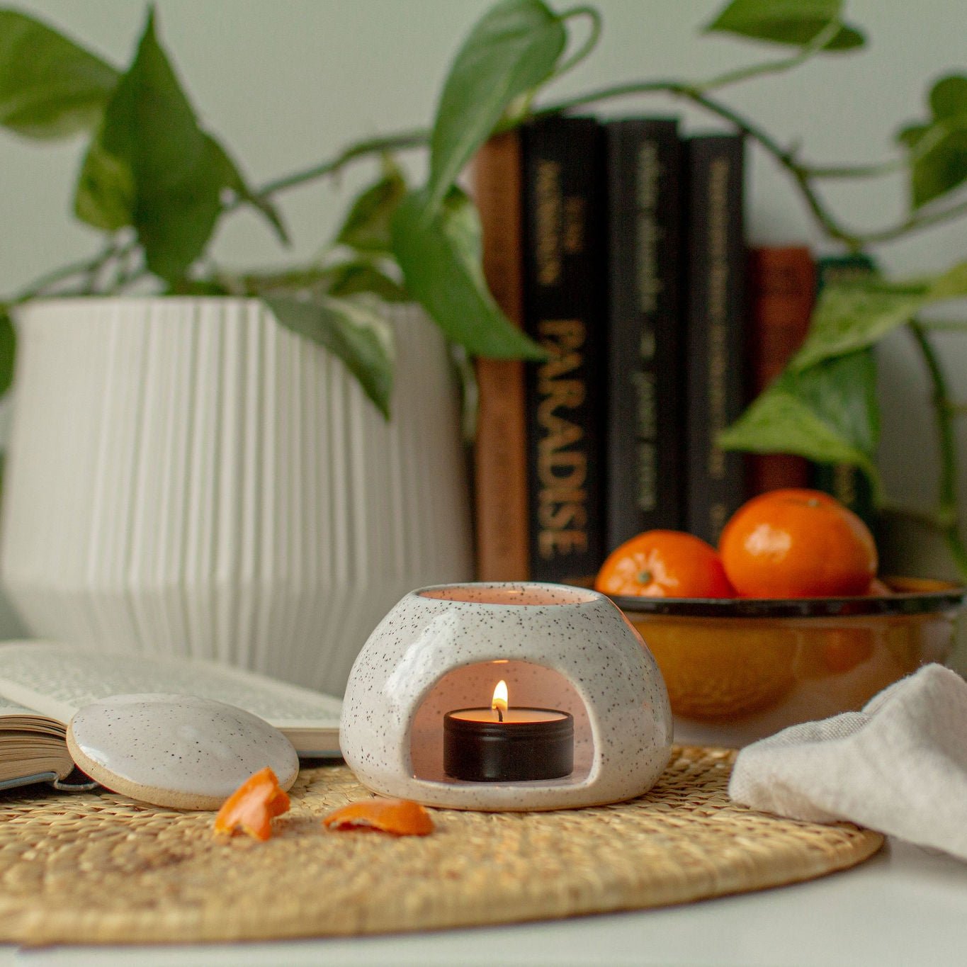 Photo of a minimalist styled ceramic tealight holder that looks like a speckled hut with a removeable lid. There's a black tin tealight burning inside the holder. It looks very cozy, set on a kitchen counter, with a green plant, some fresh mandarin oranges and books in the background, and a wicker placemat underneath the ceramic tealight house.