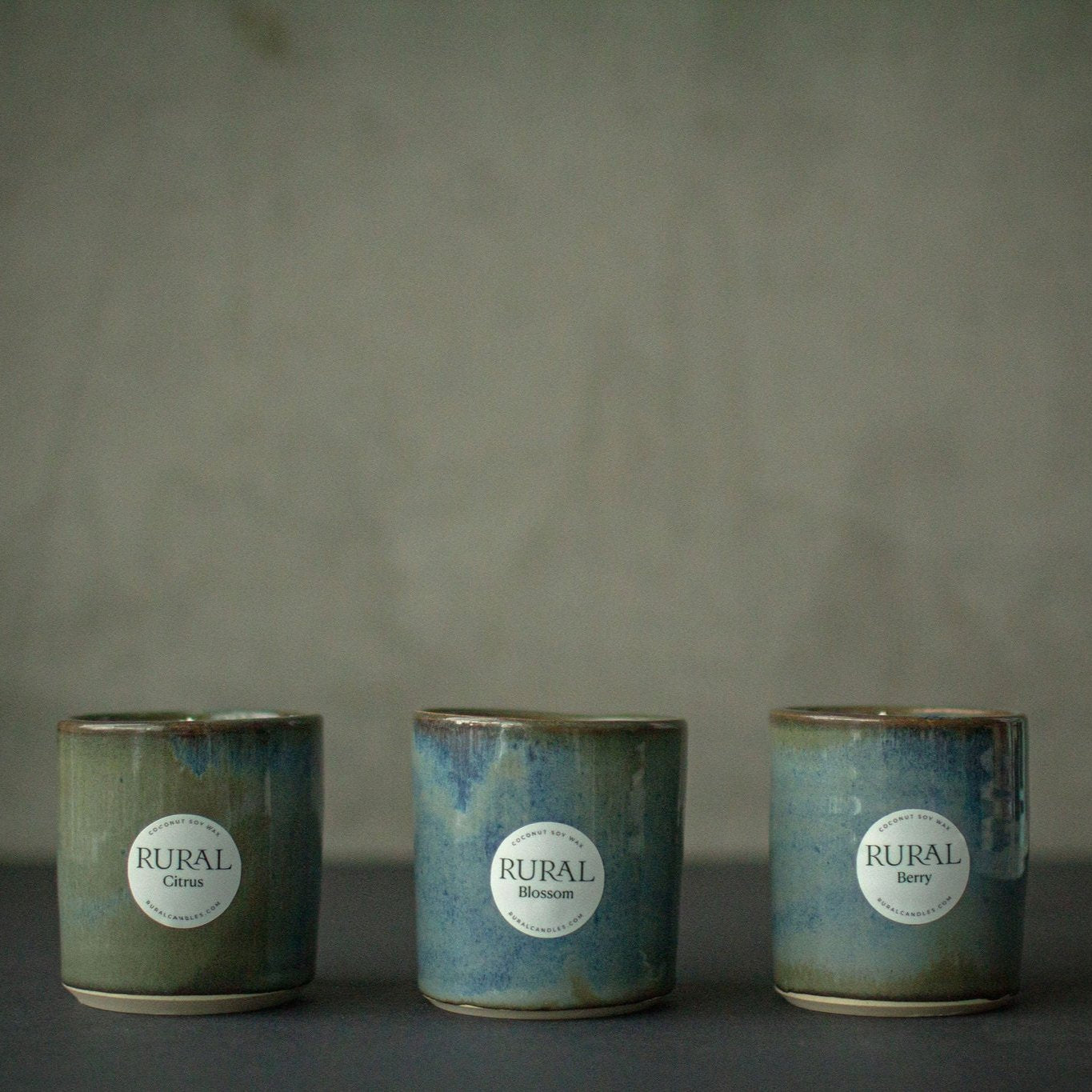 Photo of a group of three handcrafted pottery candles with a beautiful blue green and brown glaze, with white round RURAL Candles labels indicating Berry, Blossom and Citrus fragrances.
