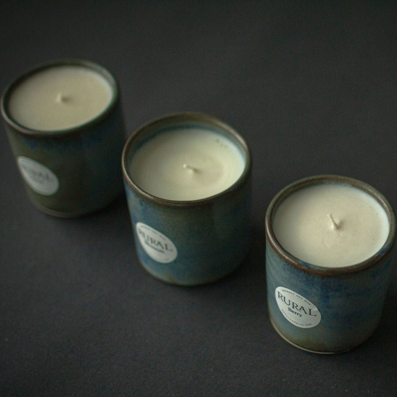 Photo of a group of three handcrafted pottery candles with a beautiful blue green and brown glaze, with white round RURAL Candles labels indicating Berry and Vacation fragrances.
